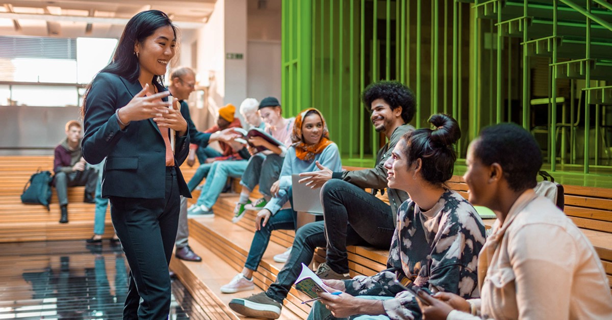 Image of a woman talking to some students in a place that could be a college or university. 