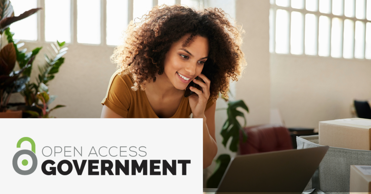 Image of a woman using her phone while consulting information on the screen of a laptop. On the lower left corner is placed the Open Access Government logo.  